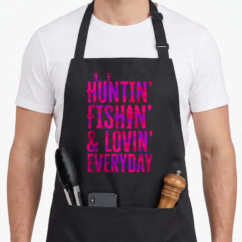 Fish camo apron - Pink purple camo text HUNTIN FISHIN LOVIN EVERYDAY with antlers and fish silhouette — shown on a black cotton canvas apron against a white background