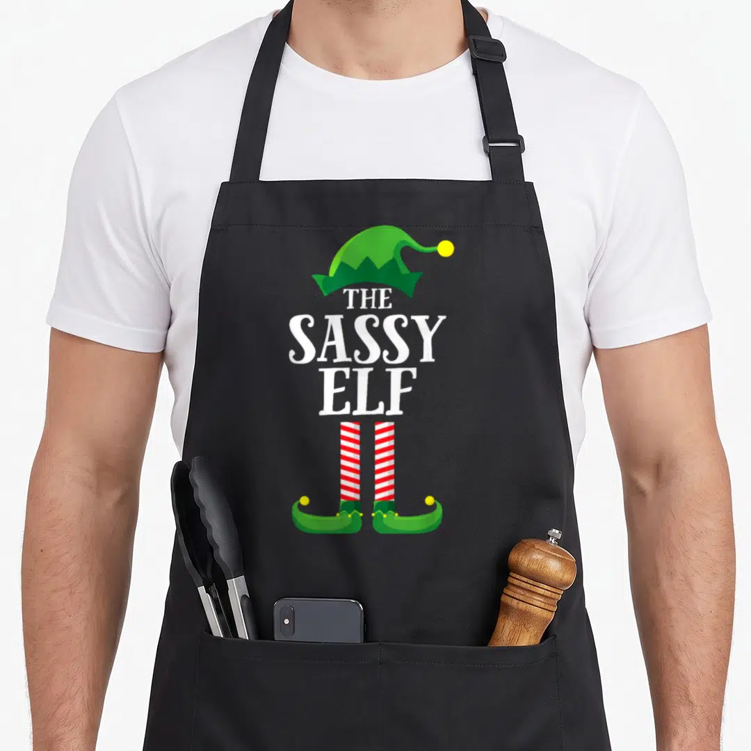 Sassy elf apron - Stacked white text between green hat and red striped legs with yellow pom-poms — shown on a black cotton canvas apron against a white background
