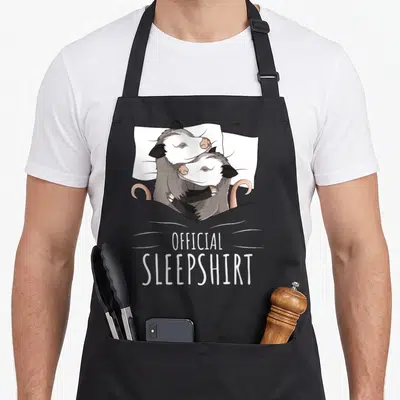 Possum novelty apron - Two grey and white opossums hugging in bed with pink tails and official sleepshirt text below — shown on a black cotton canvas apron against a white background