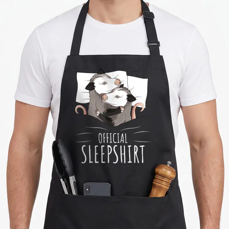 Possum novelty apron - Two grey and white opossums hugging in bed with pink tails and official sleepshirt text below — shown on a black cotton canvas apron against a white background