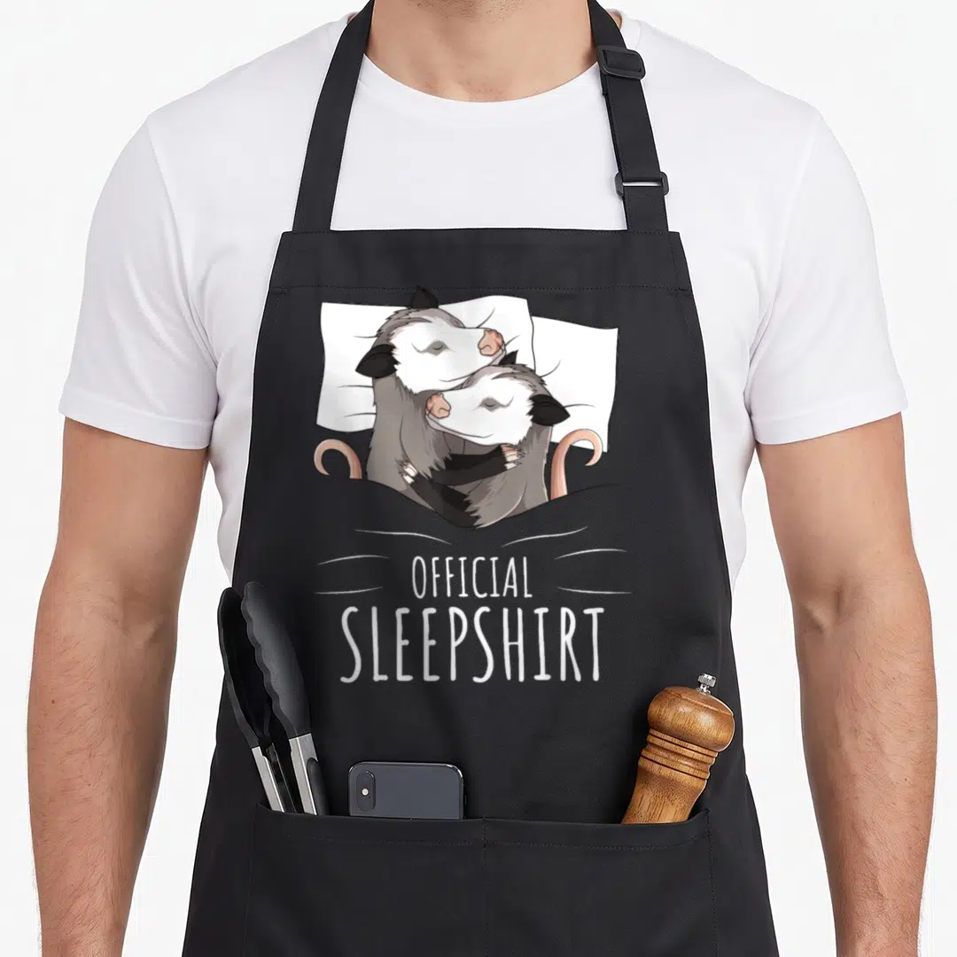 Possum novelty apron - Two grey and white opossums hugging in bed with pink tails and official sleepshirt text below — shown on a black cotton canvas apron against a white background