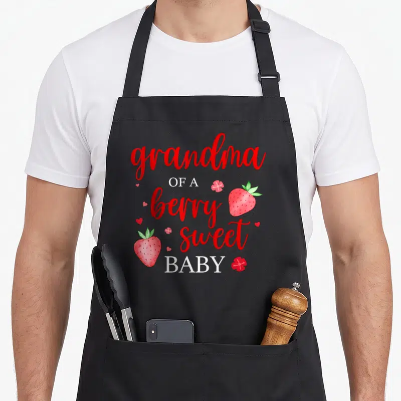 Berry grandma apron - Grandma of a berry sweet baby strawberry pun text with watercolor fruit art — shown on a black cotton canvas apron against a white background