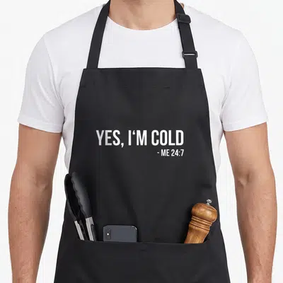 Cold winter apron - White stacked text YES I'M COLD ME 24:7 on black background — shown on a black cotton canvas apron against a white background