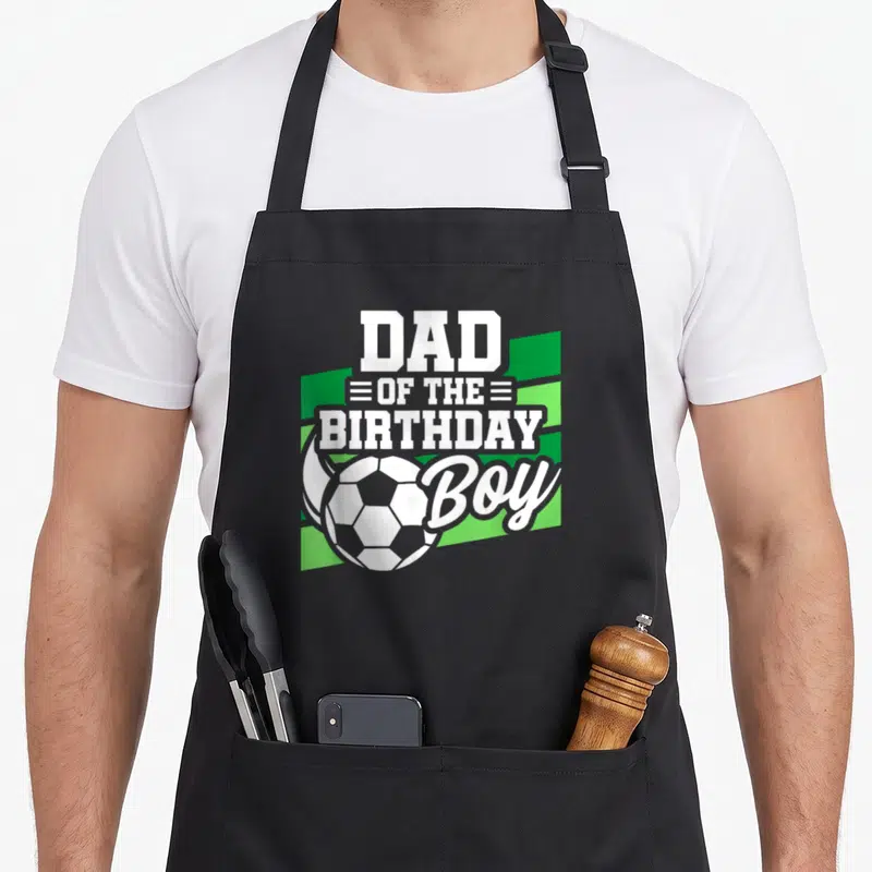 Cute soccer birthday apron - Dad of the birthday boy text with soccer ball on green stripes — shown on a black cotton canvas apron against a white background