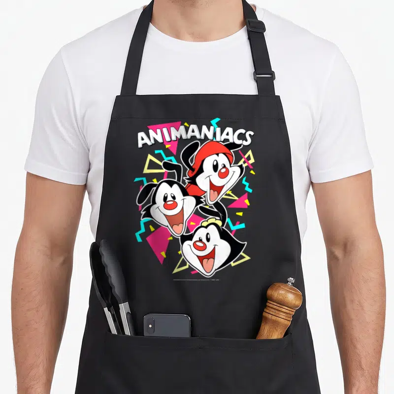 Animaniac dot apron - ANIMANIACS text above three cartoon heads with red pink yellow cyan geometric shapes — shown on a black cotton canvas apron against a white background