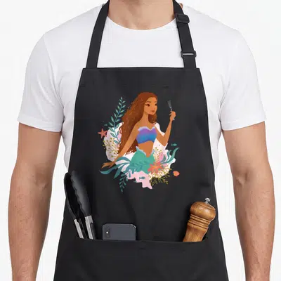 Undersea mermaid apron - Cartoon mermaid holding fork surrounded by teal seaweed, orange coral, and pink starfish — shown on a black cotton canvas apron against a white background