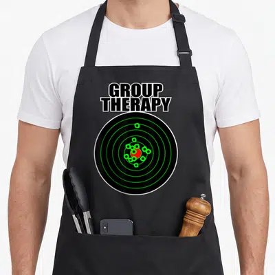 Funny my therapy apron - Stacked GROUP THERAPY text above a black target with green rings and red center — shown on a black cotton canvas apron against a white background