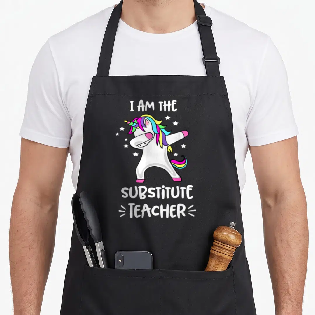 Substitute dabbing teacher apron - Cartoon unicorn dabbing with rainbow mane surrounded by stars and text — shown on a black cotton canvas apron against a white background