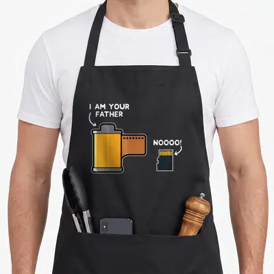Film photography apron - Yellow film canister labeled I AM YOUR FATHER points to black SD card saying NOOOO! — shown on a black cotton canvas apron against a white background