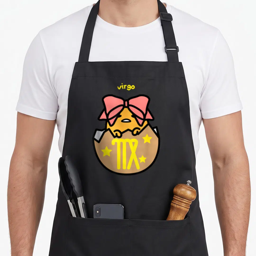 Virgo gudetama apron - Yellow egg character with pink bow hatching from tan shell with virgo text and stars — shown on a black cotton canvas apron against a white background