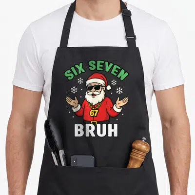 Six seven christmas apron - Cartoon Santa with sunglasses 67 chain SIX SEVEN BRUH text snowflakes red green white yellow — shown on a black cotton canvas apron against a white background