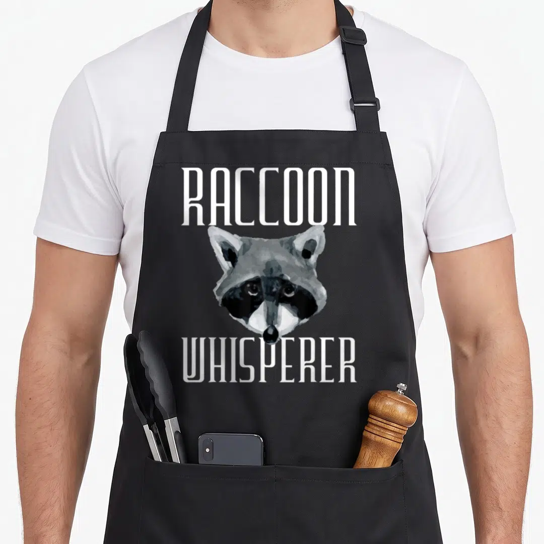 Raccoon whisperer animal apron - Realistic raccoon head centered between white RACCOON and WHISPERER text in grey — shown on a black cotton canvas apron against a white background
