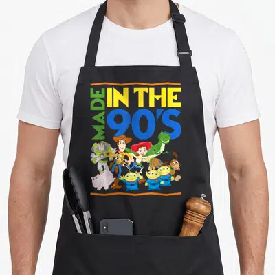 Funny shot 90s apron - Toy Story characters clustered under stacked Made In The 90's text in blue yellow green and red — shown on a black cotton canvas apron against a white background