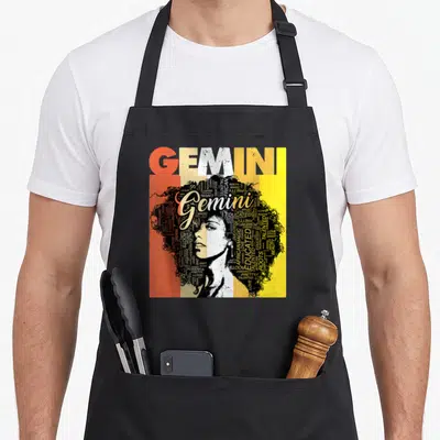 Gemini pride hair apron - Typographic woman portrait with afro made of words GEMINI ROOTS MAGIC CURRY in orange beige — shown on a black cotton canvas apron against a white background
