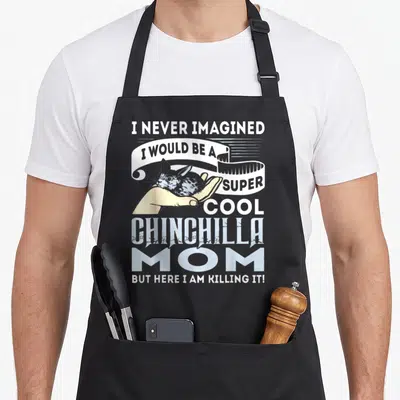 Imagined chinchilla mom apron - Distressed text I NEVER IMAGINED I WOULD BE A SUPER COOL CHINCHILLA MOM around — shown on a black cotton canvas apron against a white background