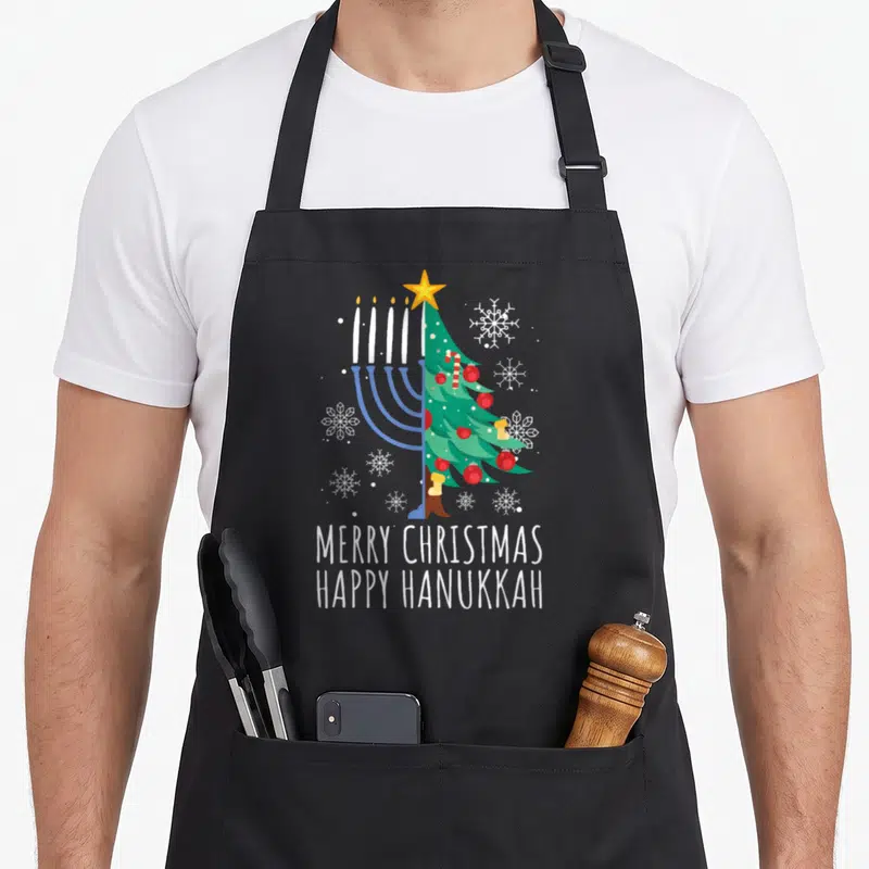 Merry hanukkah apron - Split menorah and Christmas tree graphic with white stacked text MERRY CHRISTMAS HAPPY HANUKKAH — shown on a black cotton canvas apron against a white background