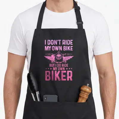 Wife biker apron - Pink winged motorcycle between I Don't Ride My Own Bike But I Do Ride My Own Biker text with six — shown on a black cotton canvas apron against a white background