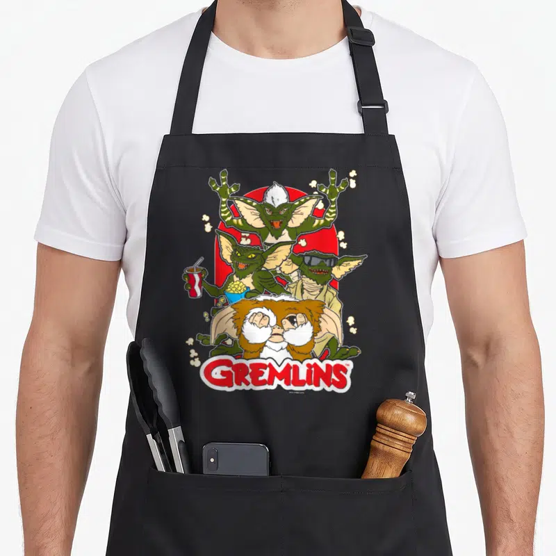 Popcorn may look apron - Four Gremlins stacked vertically on red circle with GREMLINS text at bottom — shown on a black cotton canvas apron against a white background