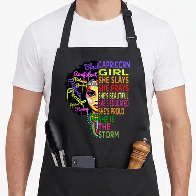 Retro capricorn birthday apron - Colorful cartoon woman face with stacked text CAPRICORN GIRL SHE SLAYS SHE IS — shown on a black cotton canvas apron against a white background