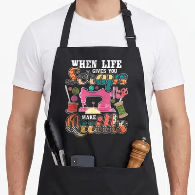 Scrap quilt apron - Pink sewing machine surrounded by buttons and spools under patchwork text reading When Life Gives — shown on a black cotton canvas apron against a white background