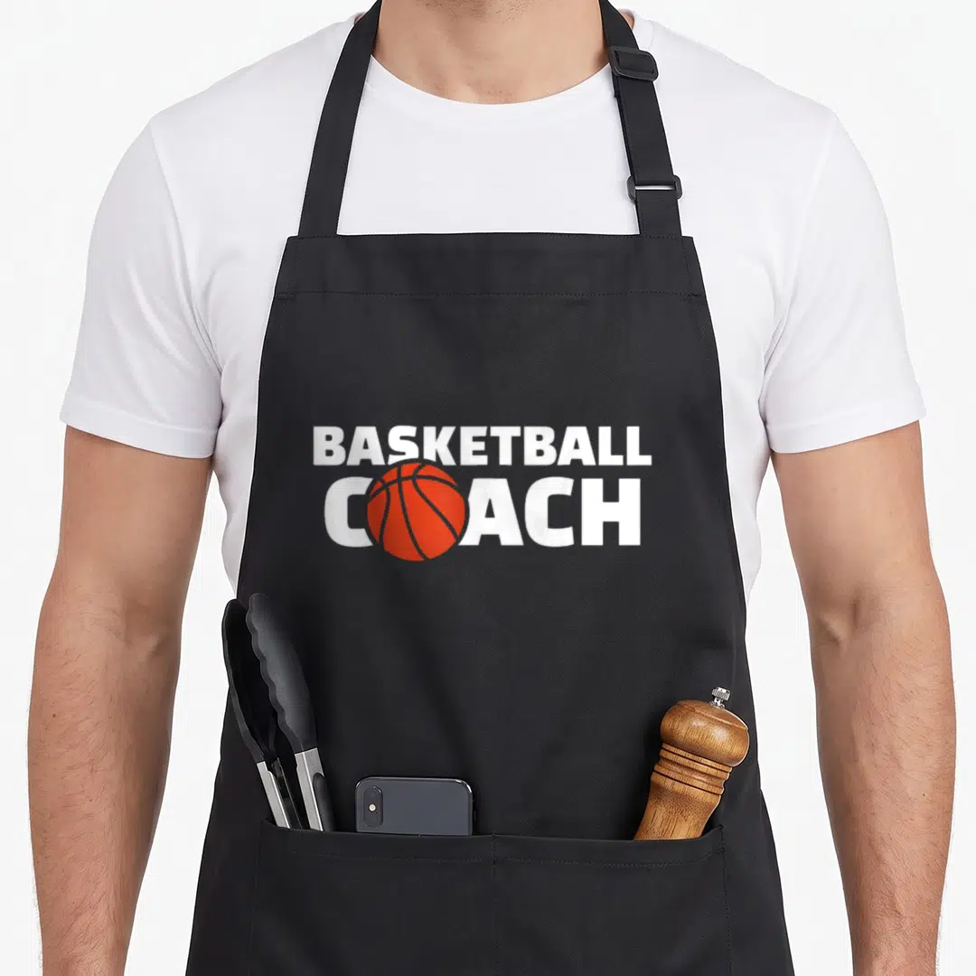 Cute basketball coach apron - White BASKETBALL COACH text with orange basketball replacing the O on a black background — shown on a black cotton canvas apron against a white background