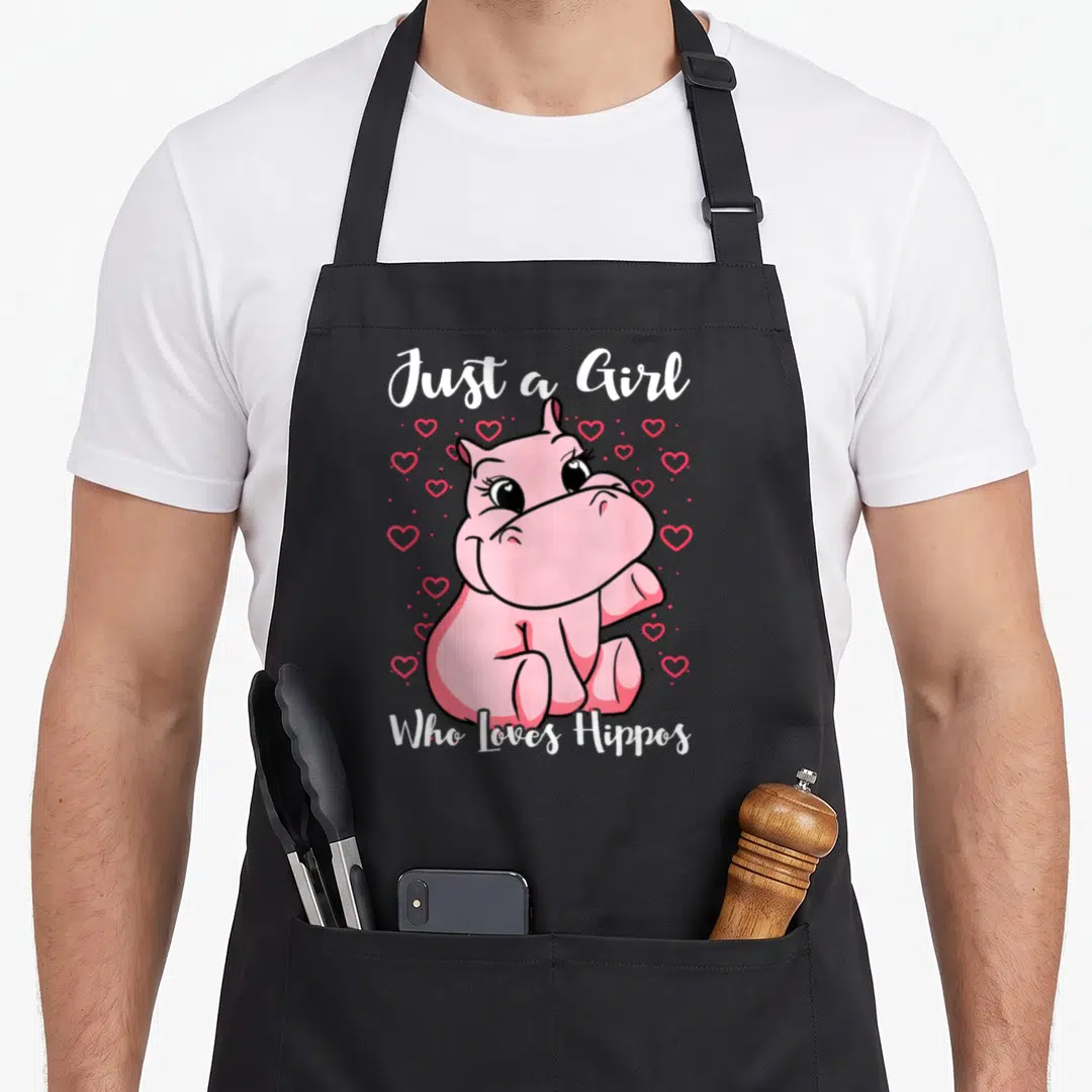 Funny hippo apron - Pink cartoon hippo sitting surrounded by red hearts with white text Just a Girl Who Loves Hippos — shown on a black cotton canvas apron against a white background