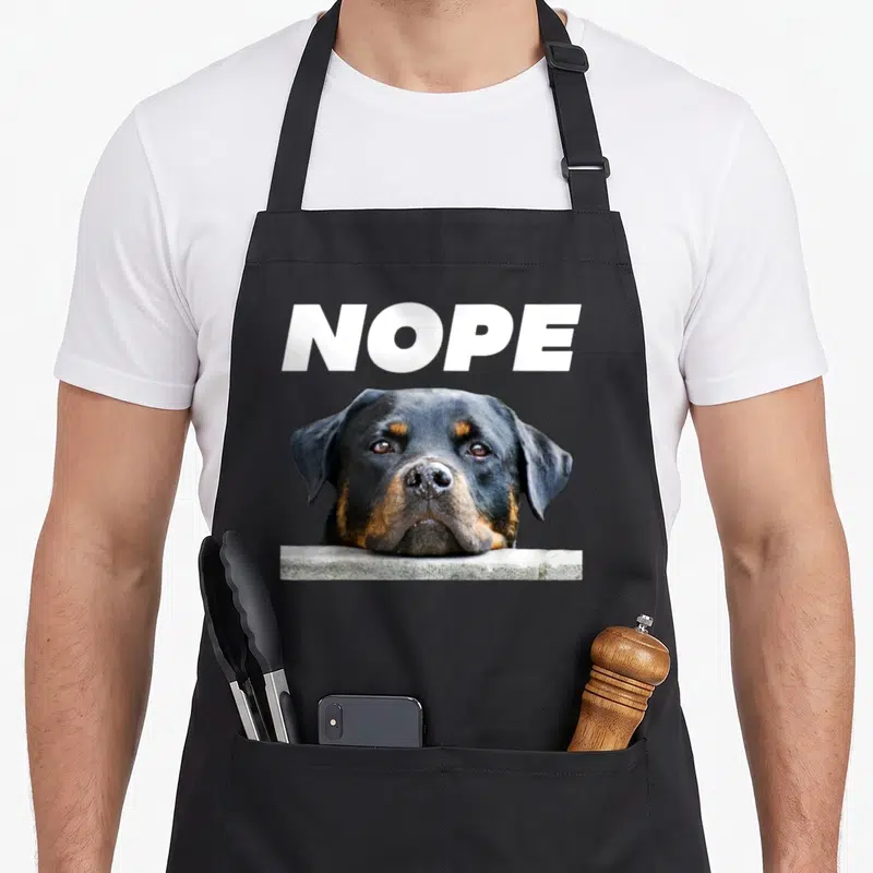 Rotweiller dog face apron - White NOPE text above a black tan dog chin resting on a grey ledge — shown on a black cotton canvas apron against a white background