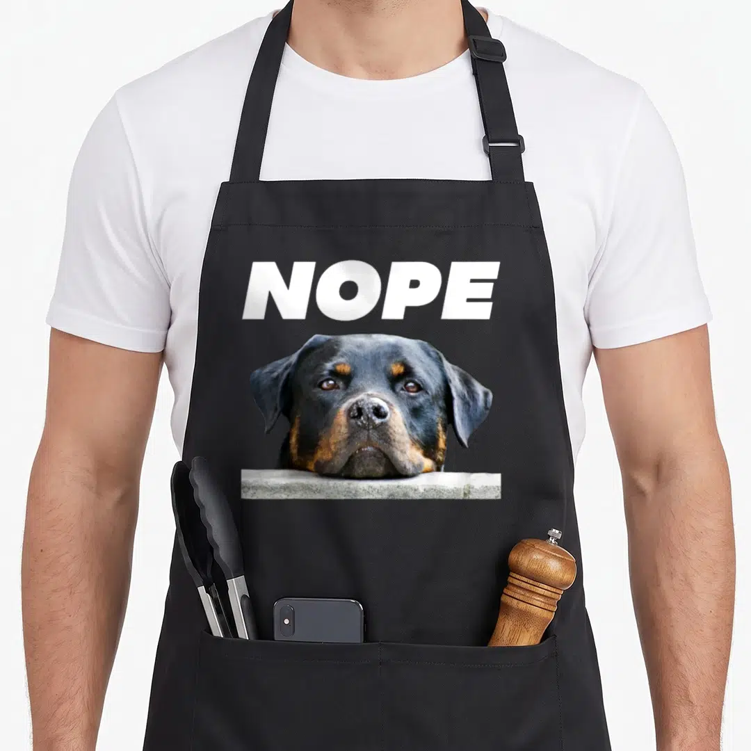 Rotweiller dog face apron - White NOPE text above a black tan dog chin resting on a grey ledge — shown on a black cotton canvas apron against a white background