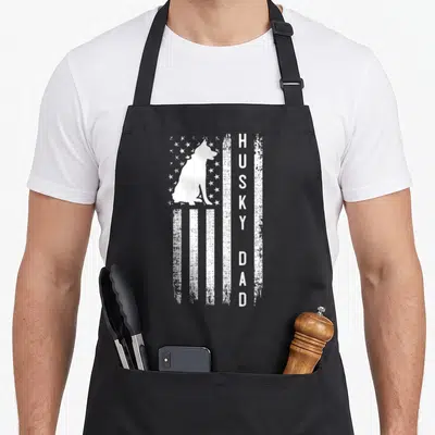 Siberian top dog aprons - White husky silhouette and vertical HUSKY DAD text on distressed flag pattern — shown on a black cotton canvas apron against a white background