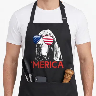 Basset 4th of july apron - White basset hound head with flag sunglasses and red merica text below — shown on a black cotton canvas apron against a white background