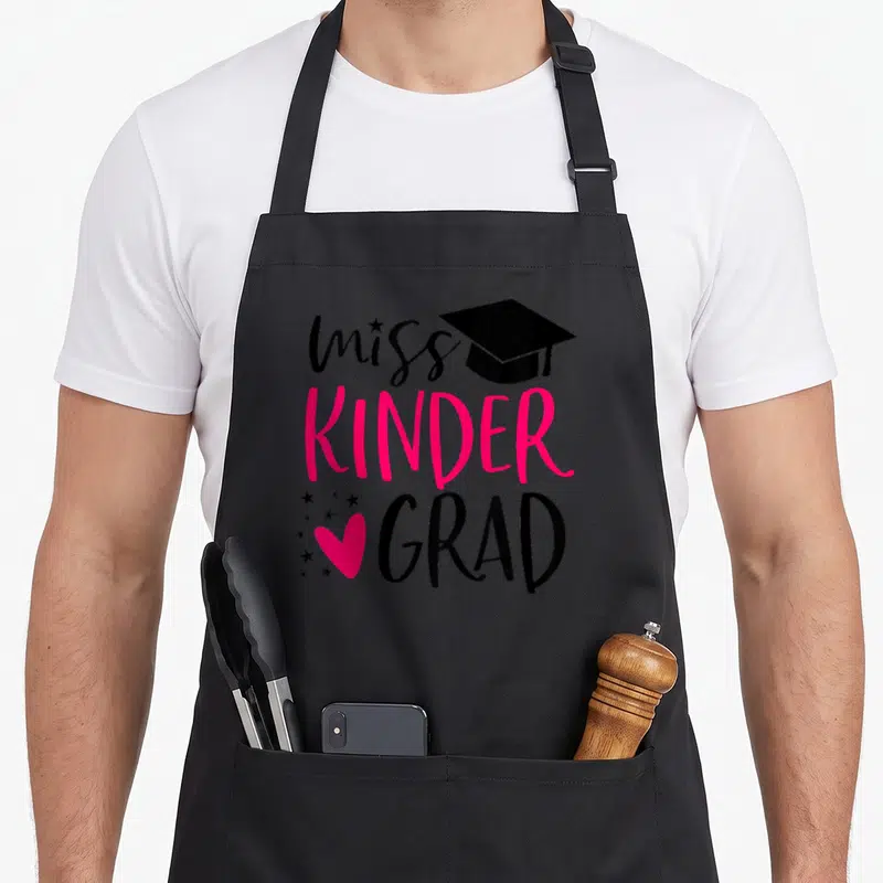 Miss kindergarten apron - Black graduation cap next to miss text with pink KINDER GRAD and pink heart plus black stars — shown on a black cotton canvas apron against a white background