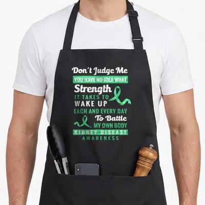 Judge kidney disease awareness apron - Distressed white and green text with two green awareness ribbons reading Don't — shown on a black cotton canvas apron against a white background