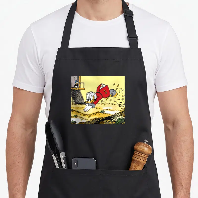 Ducktale cash apron - Scrooge McDuck diving into coins and bills with Donald watching from a platform — shown on a black cotton canvas apron against a white background