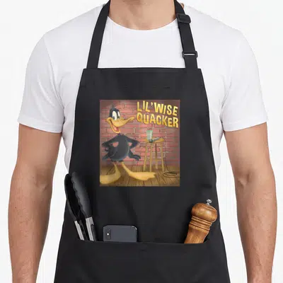 Lil duck apron - Daffy Duck standing on wooden floor near stool with mic and glass against red brick wall — shown on a black cotton canvas apron against a white background