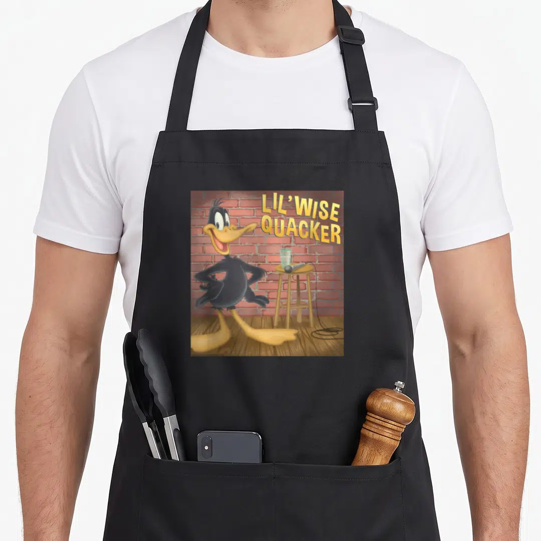 Lil duck apron - Daffy Duck standing on wooden floor near stool with mic and glass against red brick wall — shown on a black cotton canvas apron against a white background