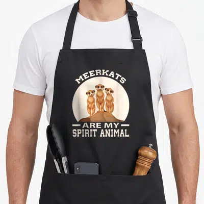 Meerkat animal apron - Three cartoon meerkats on rock inside beige circle with arched and stacked white text — shown on a black cotton canvas apron against a white background