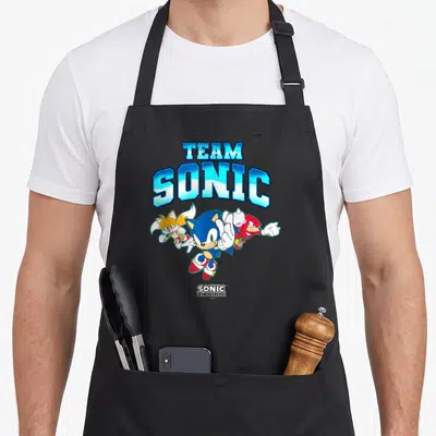 Main crazy hedgehog man apron - TEAM SONIC text above three cartoon hedgehogs and fox in blue red orange — shown on a black cotton canvas apron against a white background