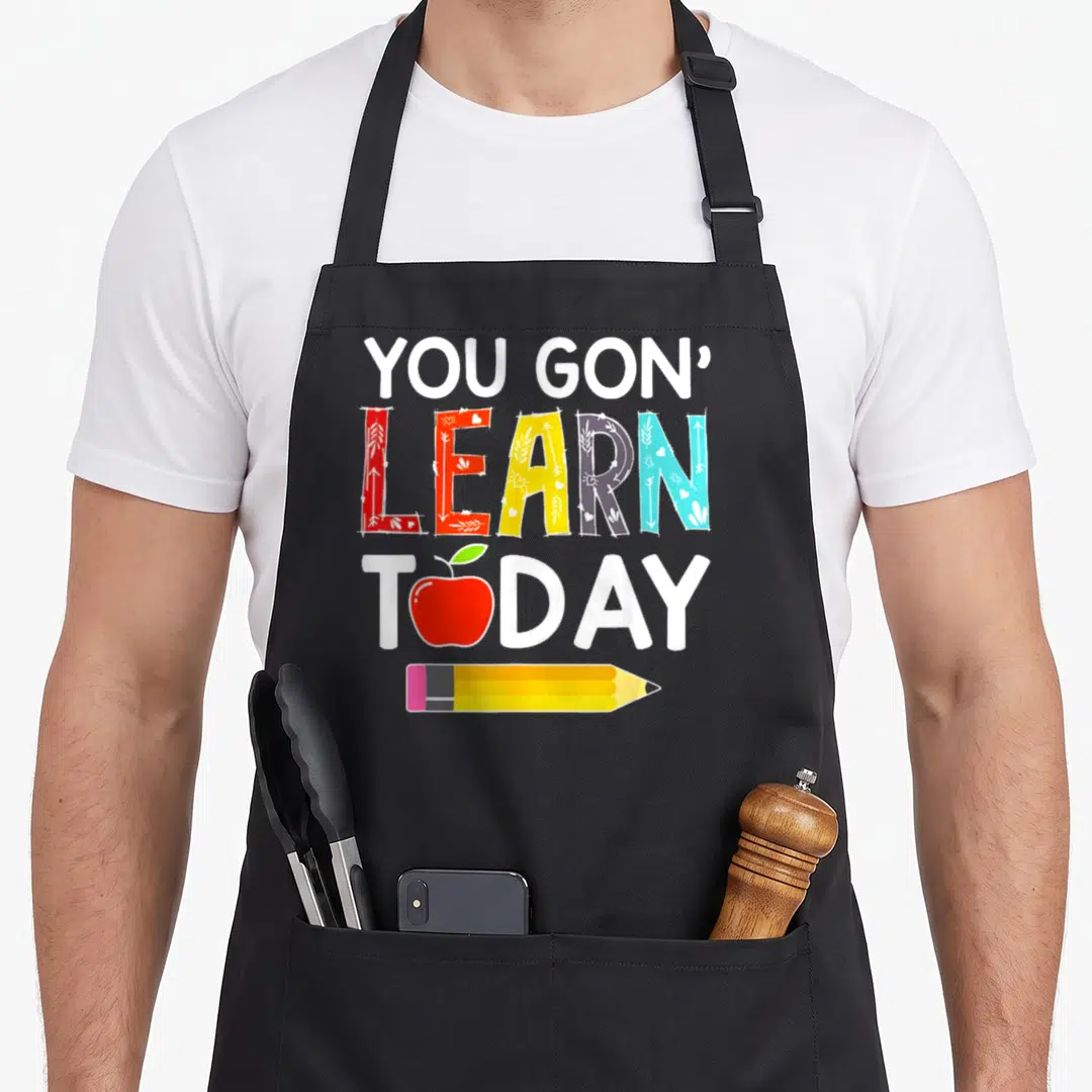 Gon teacher apron - Stacked YOU GON LEARN TODAY text with colorful patterned letters, red apple, and yellow pencil — shown on a black cotton canvas apron against a white background