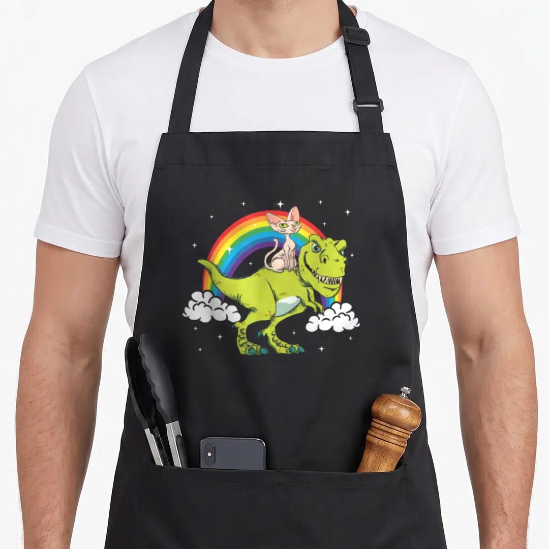 Dinosaur cat apron - Green dinosaur and beige cat on rainbow with white stars and clouds — shown on a black cotton canvas apron against a white background