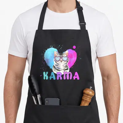 Karma cat apron - Tabby cat wearing pink heart glasses in front of blue and pink watercolor heart with text — shown on a black cotton canvas apron against a white background