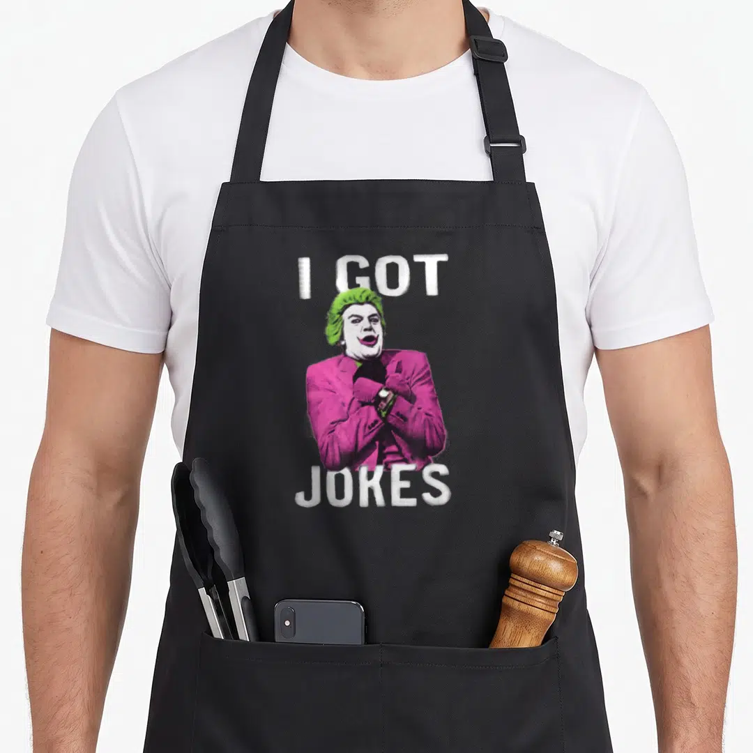 Funny joke apron - I GOT JOKES text framing a cartoon man with green hair and pink suit — shown on a black cotton canvas apron against a white background