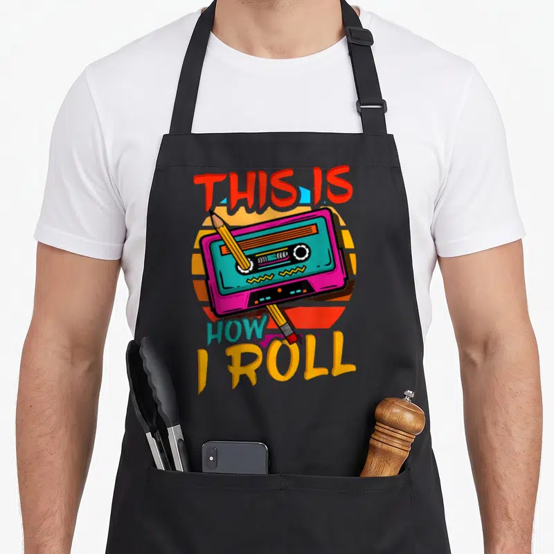 90s music 80s apron - Cassette tape with pencil crossing over retro sun, text THIS IS HOW I ROLL in red yellow pink — shown on a black cotton canvas apron against a white background