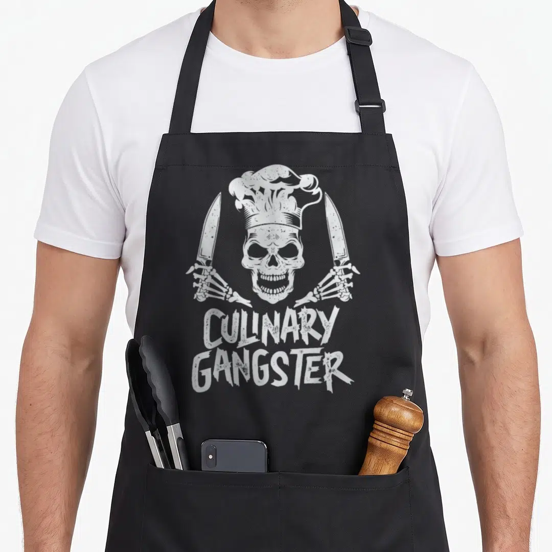 Gangster culinary aprons - White skull in chef hat holding two knives with stacked CULINARY GANGSTER text below — shown on a black cotton canvas apron against a white background