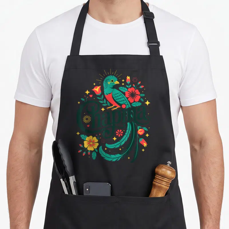 Pride guatemalan aprons - Chapina script with teal quetzal bird, red and yellow flowers, green leaves, and yellow stars — shown on a black cotton canvas apron against a white background