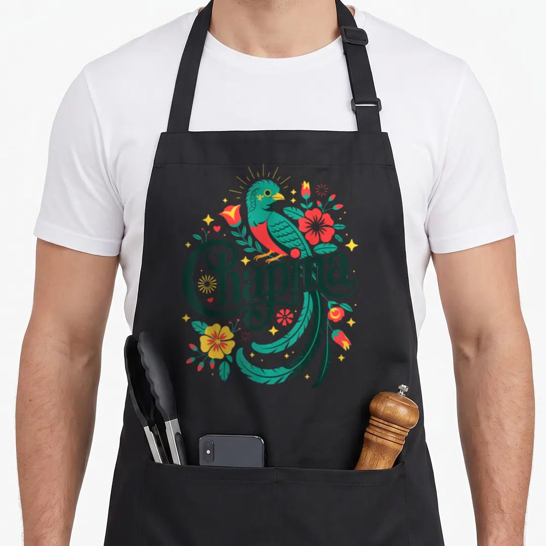Pride guatemalan aprons - Chapina script with teal quetzal bird, red and yellow flowers, green leaves, and yellow stars — shown on a black cotton canvas apron against a white background