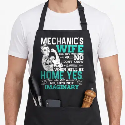 Wife auto mechanic apron - Retro woman flexing with text Yes He's Working No Shock Absorbers Two Helmets — shown on a black cotton canvas apron against a white background