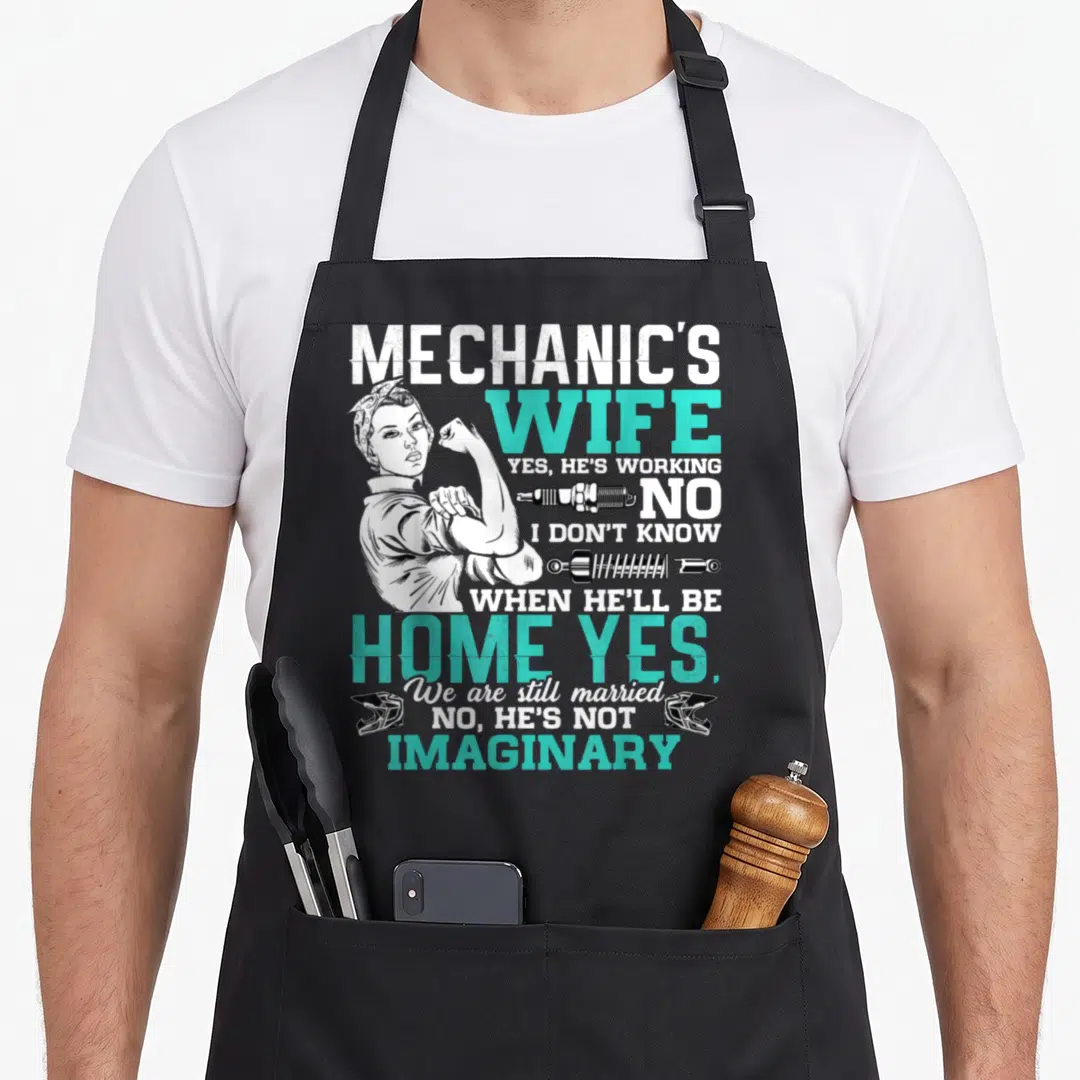 Wife auto mechanic apron - Retro woman flexing with text Yes He's Working No Shock Absorbers Two Helmets — shown on a black cotton canvas apron against a white background