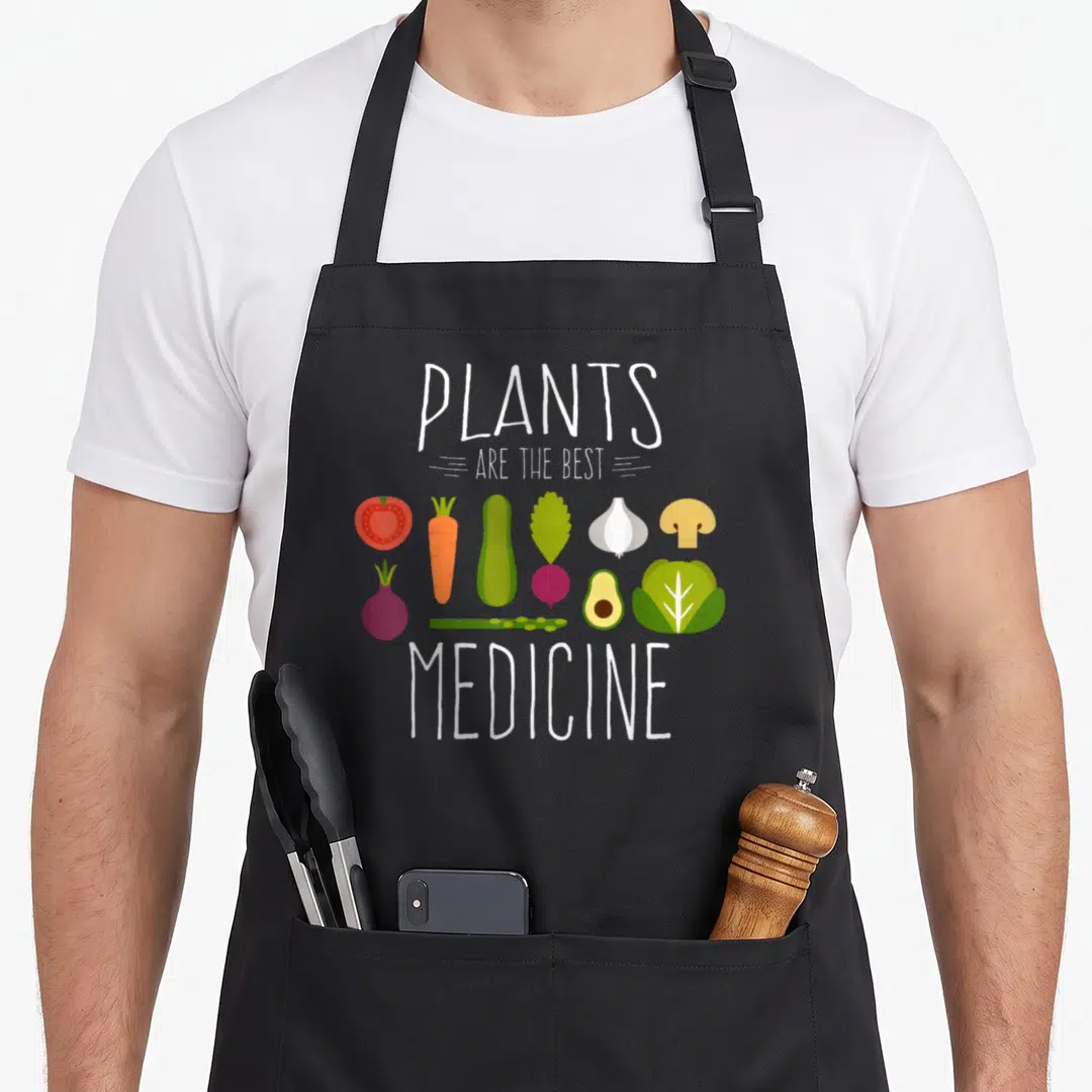 Plant vegetable apron - PLANTS ARE THE BEST MEDICINE text with cartoon vegetables in two rows — shown on a black cotton canvas apron against a white background