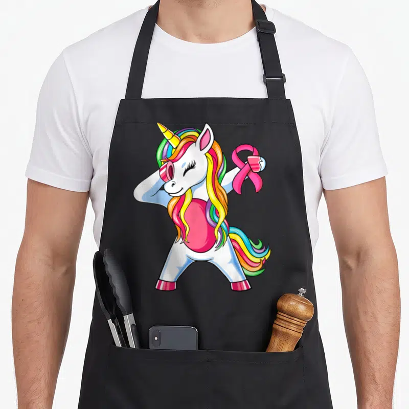Unicorn ribbon apron - Cartoon white unicorn with rainbow mane holding a pink ribbon symbol — shown on a black cotton canvas apron against a white background