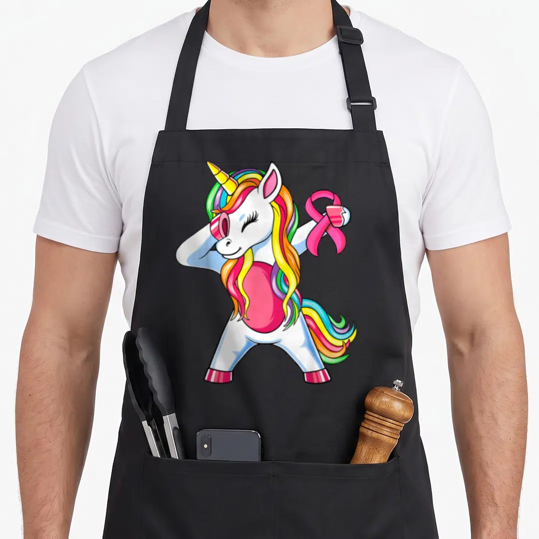 Unicorn ribbon apron - Cartoon white unicorn with rainbow mane holding a pink ribbon symbol — shown on a black cotton canvas apron against a white background